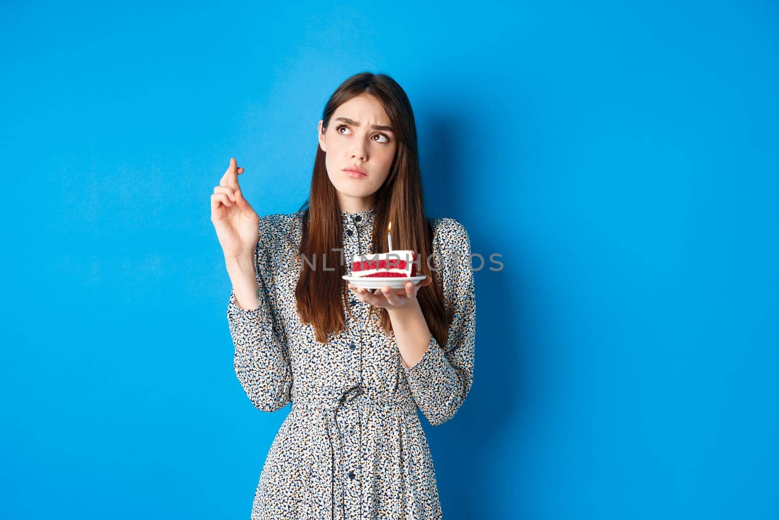 Thoughtful birthday girl making wish, cross fingers good luck, holding cake and looking at upper left corner, blue background.