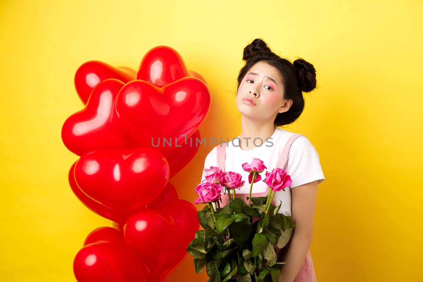 Happy Valentines concept. Sad and gloomy asian woman holding bouquet of roses and feeling upset and lonely on romantic lovers day, standing near red heart balloons, yellow background.