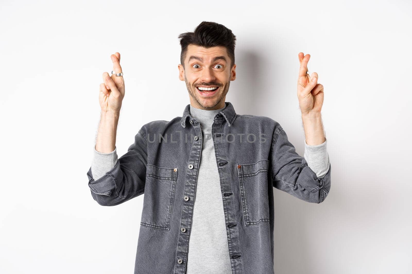 Young optimistic male hoping for dream come true, making wish with fingers crossed, smiling and expecting good news, standing on white background.