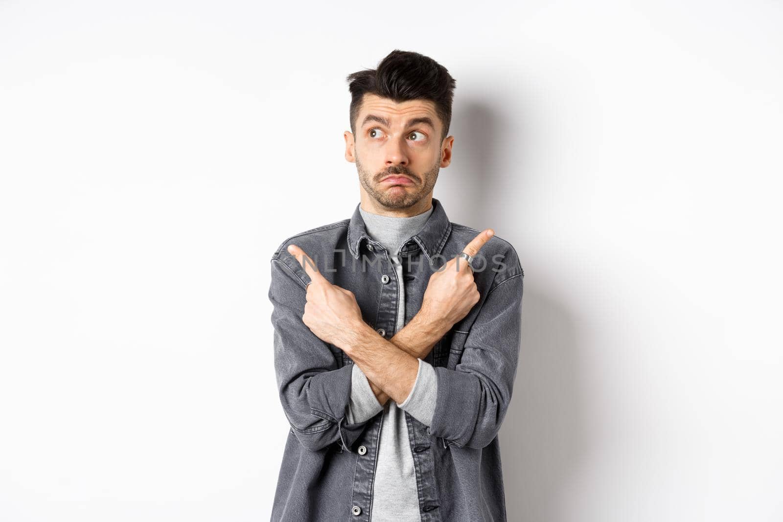 Indecisive handsome guy pointing fingers sideways, looking aside and making choice, showing two variants, standing unsure on white background.