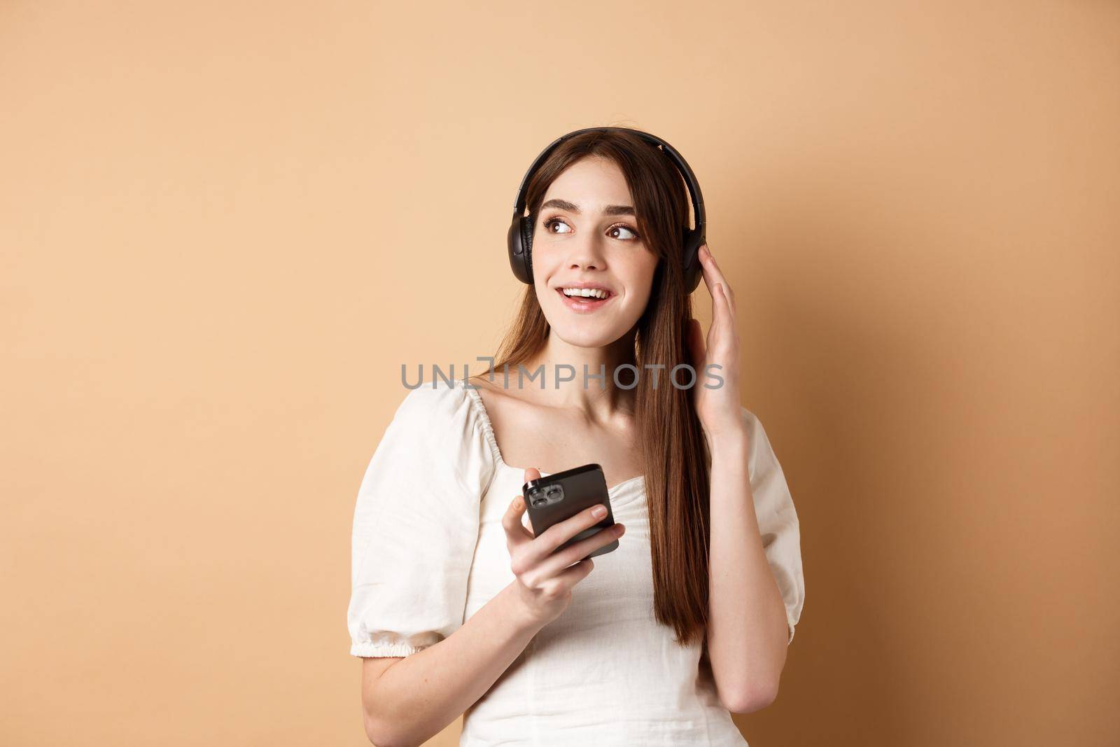 Attractive girl listening music in wireless headphones, holding mobile phone and looking aside with dreamy smile, beige background by Benzoix