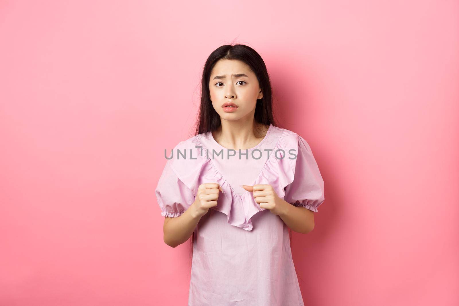 Scared teen asian girl clench fists and looking alarmed at camera, frightened with something scary, standing on pink background.