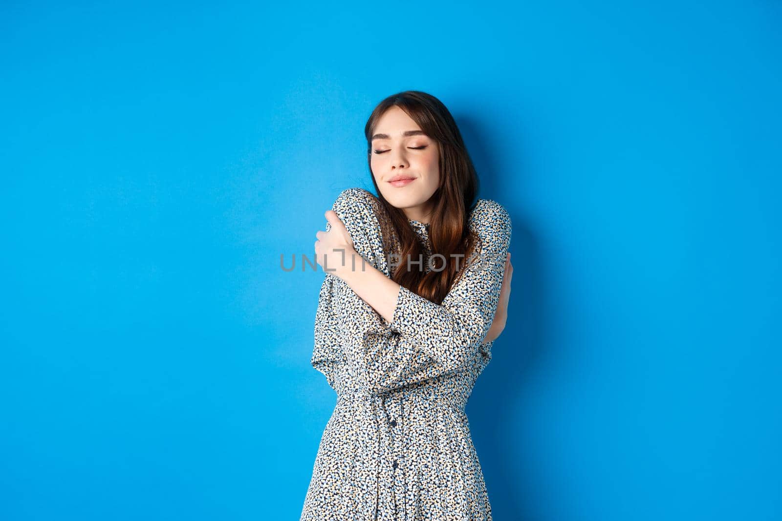 Romantic young natural woman in dress close eyes, smiling and hugging herself with love and tenderness, standing on blue background.
