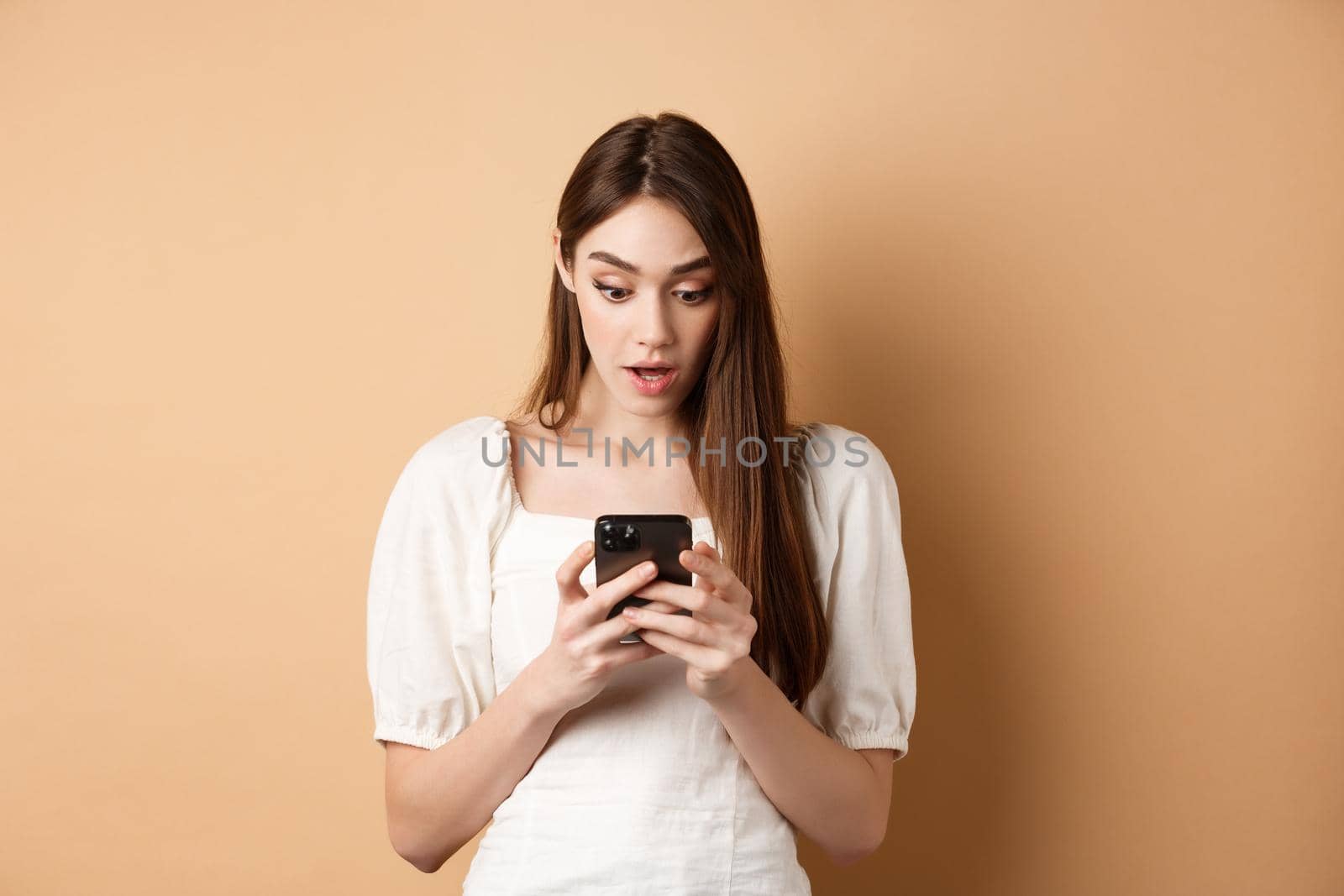 Woman look surprised at smartphone screen, reading shocking message on cell phone, standing amazed on beige background by Benzoix