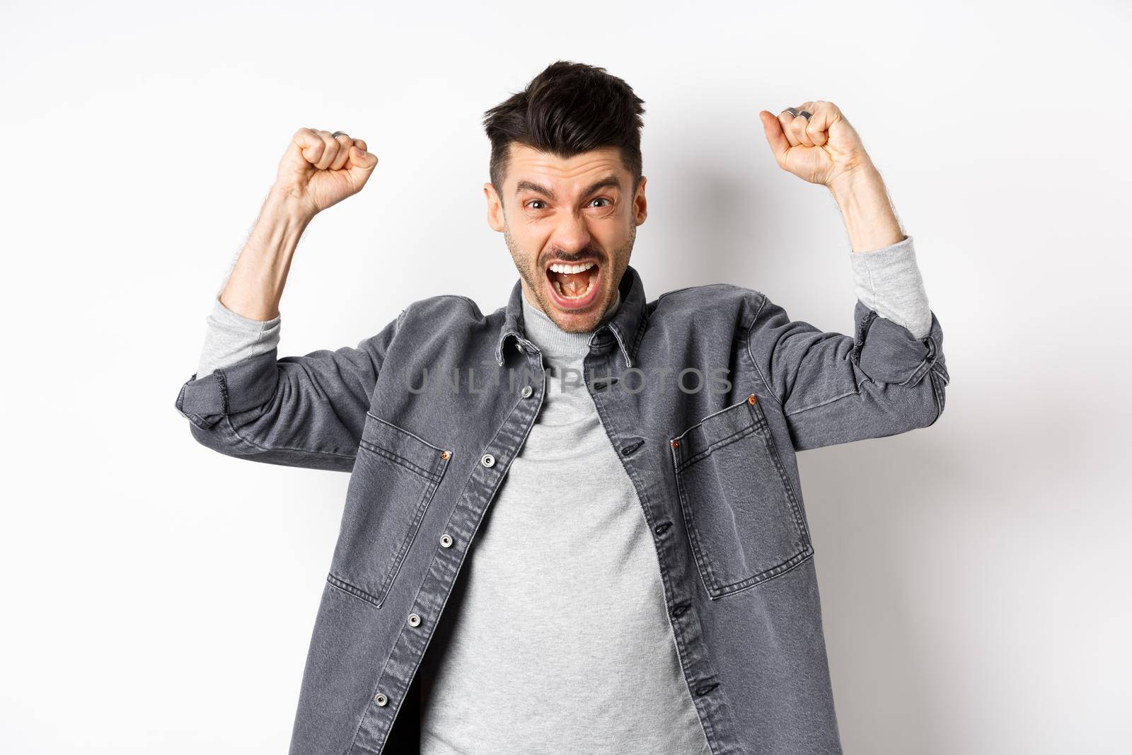 Motivated young man shouting and raising hands up, watching sports game and rooting for team, cheering and chanting, standing on white background.