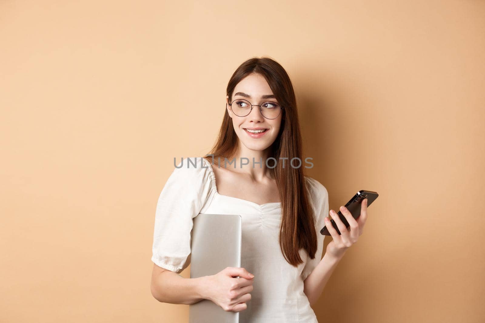 Image of young stylish woman going on work, holding laptop and smartphone, looking aside at empty space, standing on beige background by Benzoix