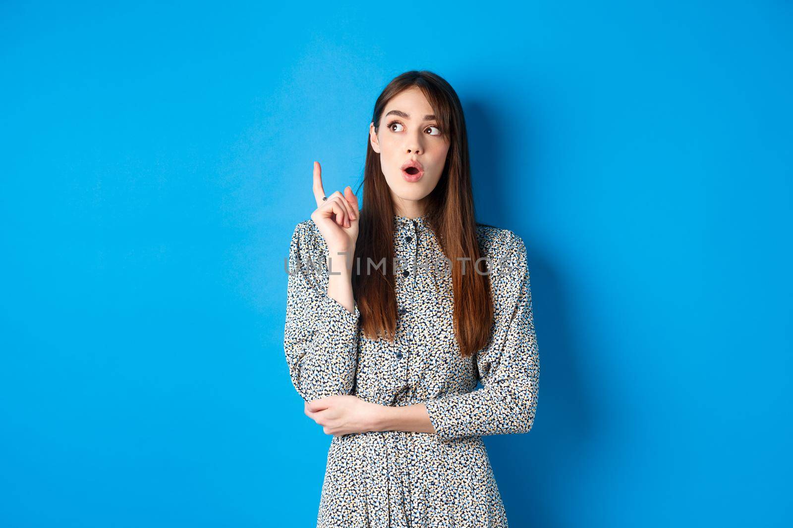 Thoughtful attractive girl in dress pitching an idea, raising finger and looking up, have a plan, standing on blue background.