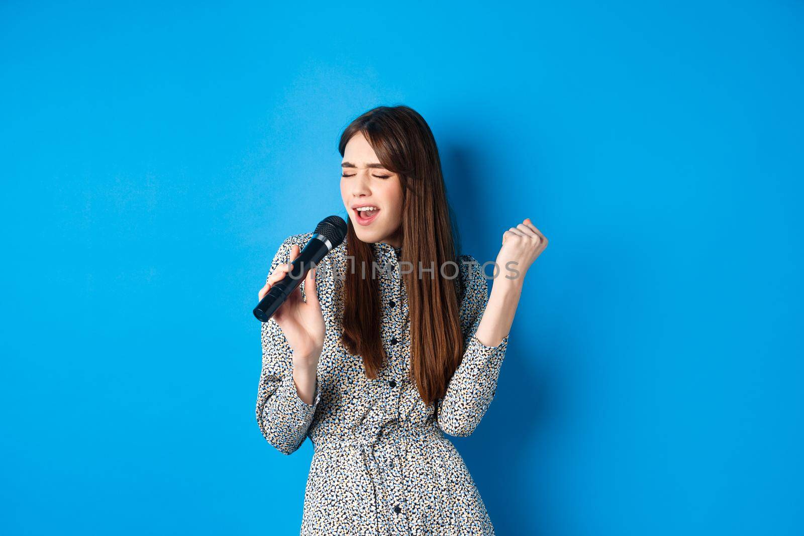 Pretty natural girl in dress singing songs in microphone, holding mic and looking passionate while performing, standing on blue background by Benzoix