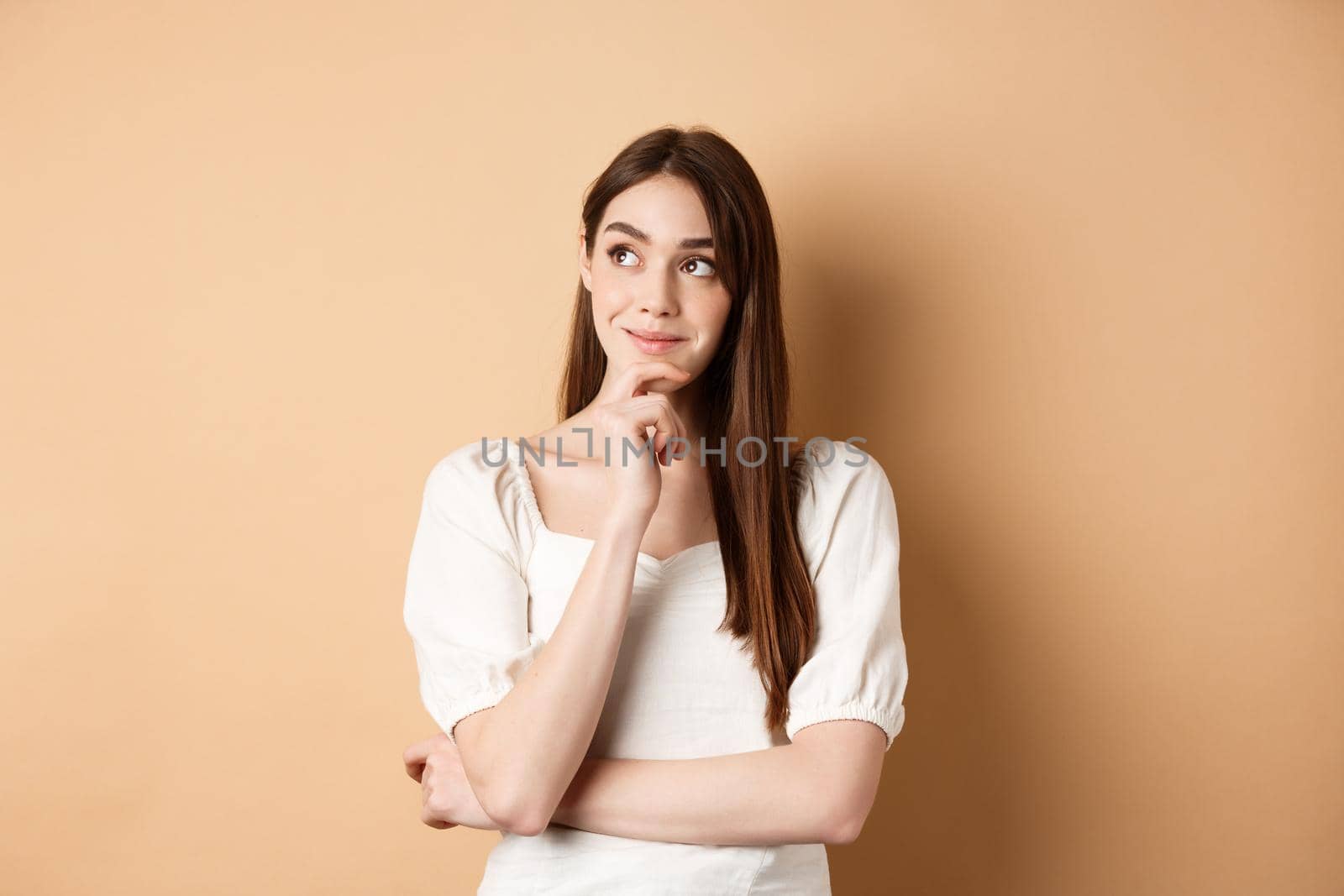 Very interesting. Pensive cute girl thinking and looking at upper left corner logo, smiling pleased, having good idea, standing on beige background.