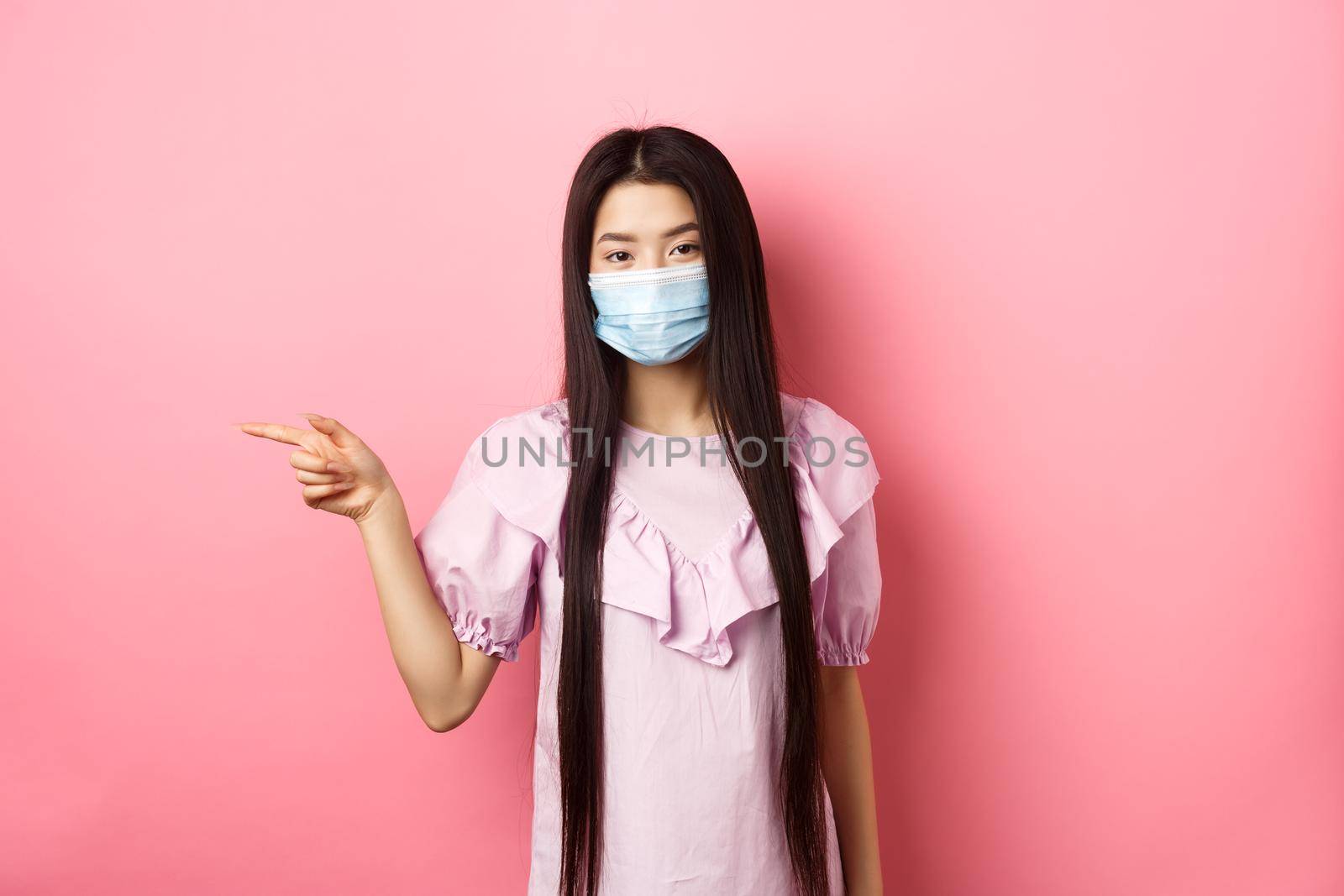 Covid-19, pandemic lifestyle concept. Cute asian teen girl in medical mask pointing finger left at logo, showing advertisement, standing in dress on pink background.