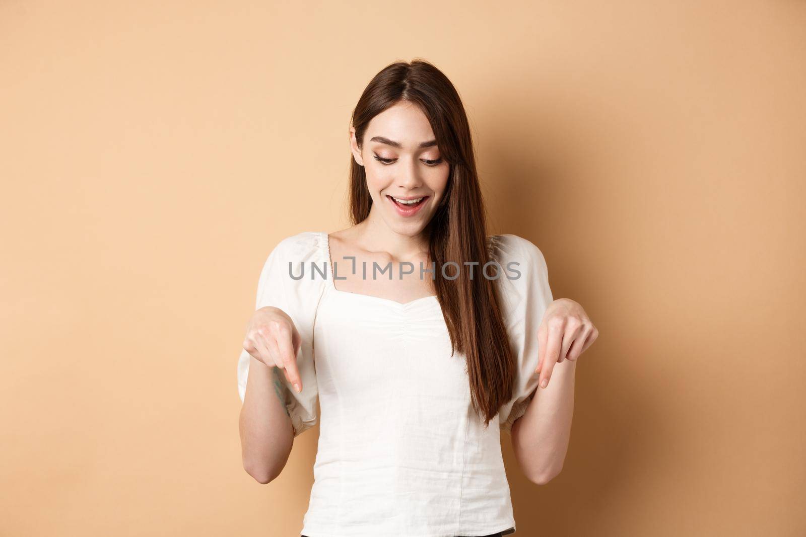 Surprised cute girl say wow, smiling amazed and pointing fingers down, showing advertisement, standing on beige background.
