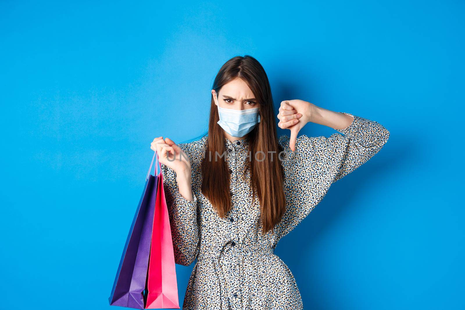 Covid-19, pandemic and lifestyle concept. Angry client in medical mask showing thumb down in dislike, frowning upset, holding shopping bags, blue background.