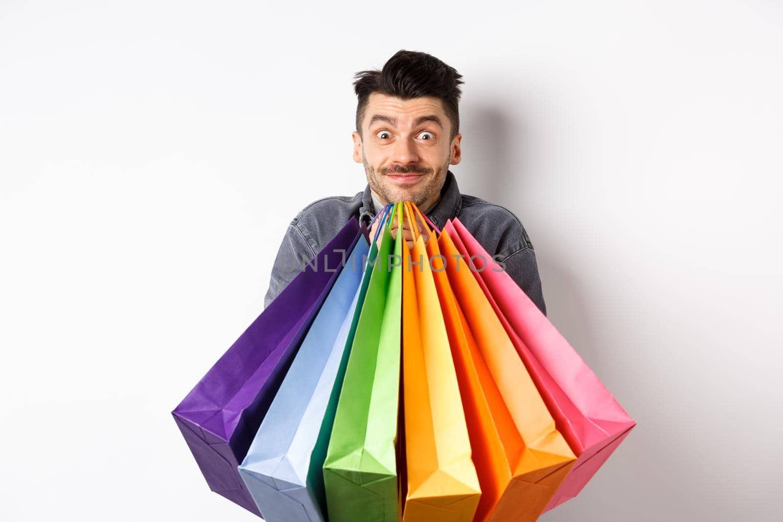 Excited young man carry colorful shopping bags and smiling happy, shopper buying on sale, standing on white background.