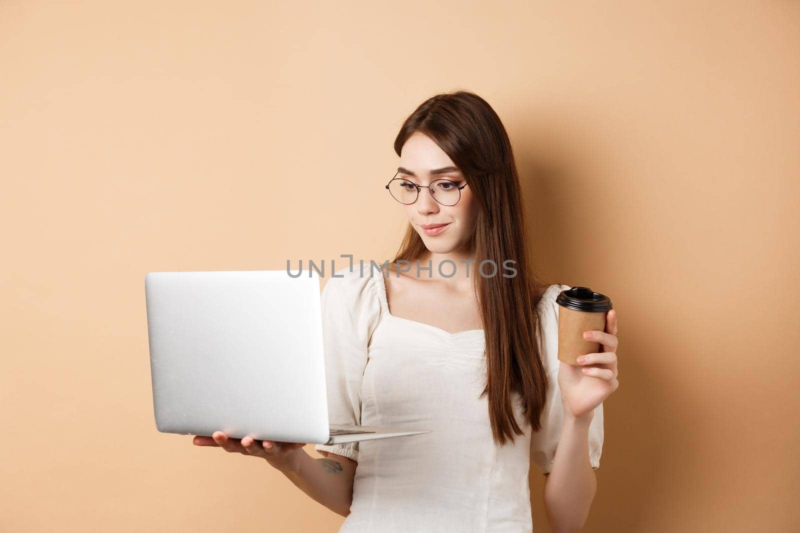 Portrait of woman working on laptop and drinking coffee. Girl student using computer for remote studies, standing on beige background.