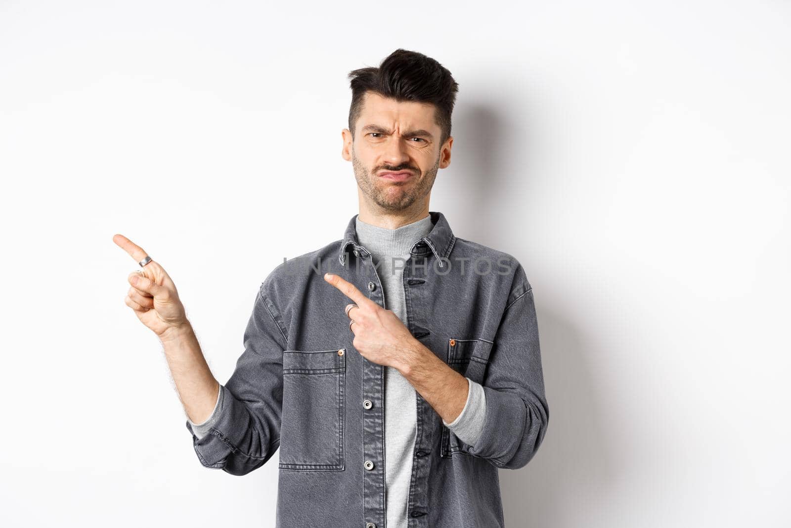 Skeptical and disappointed young man pointing fingers left, grimacing and shaking head in rejection, dislike bad product, standing on white background.