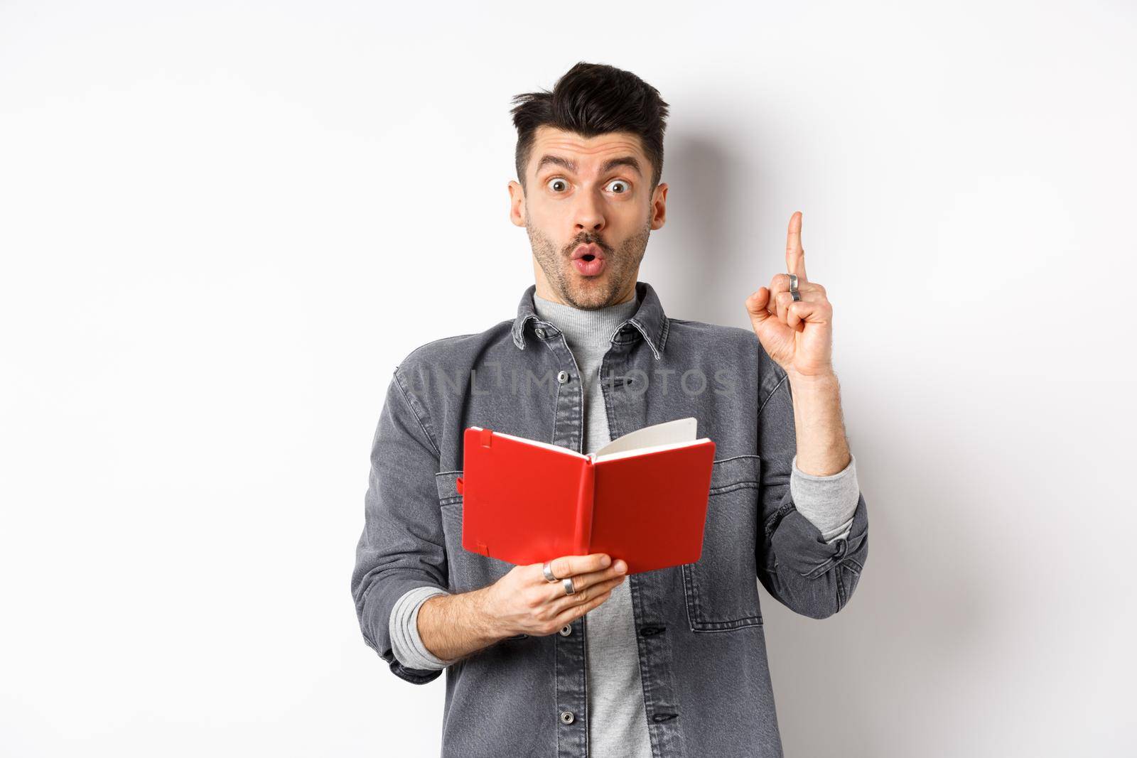 Excited man pitch an idea while reading planner or diary, holding red journal and gasping amazed, raising finger in eureka sign, standing on white background.