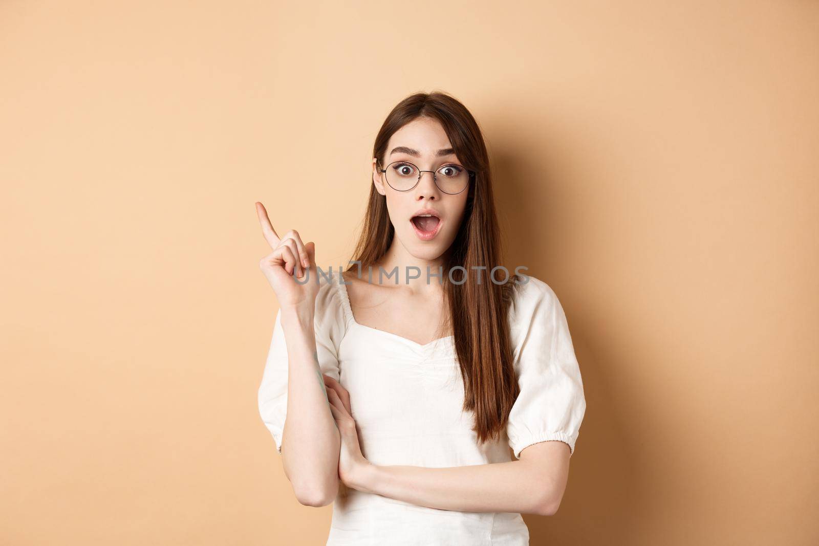 Creative young woman in glasses pitching idea, raising finger in eureka sign and gasping, have a plan, standing on beige background by Benzoix