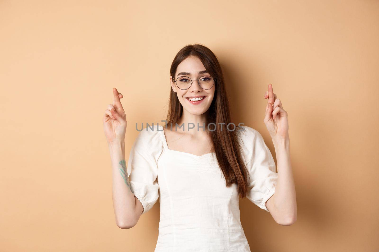 Hopeful smiling girl making wish, cross fingers for good luck and looking happy, praying for dream to come true, standing on beige background.