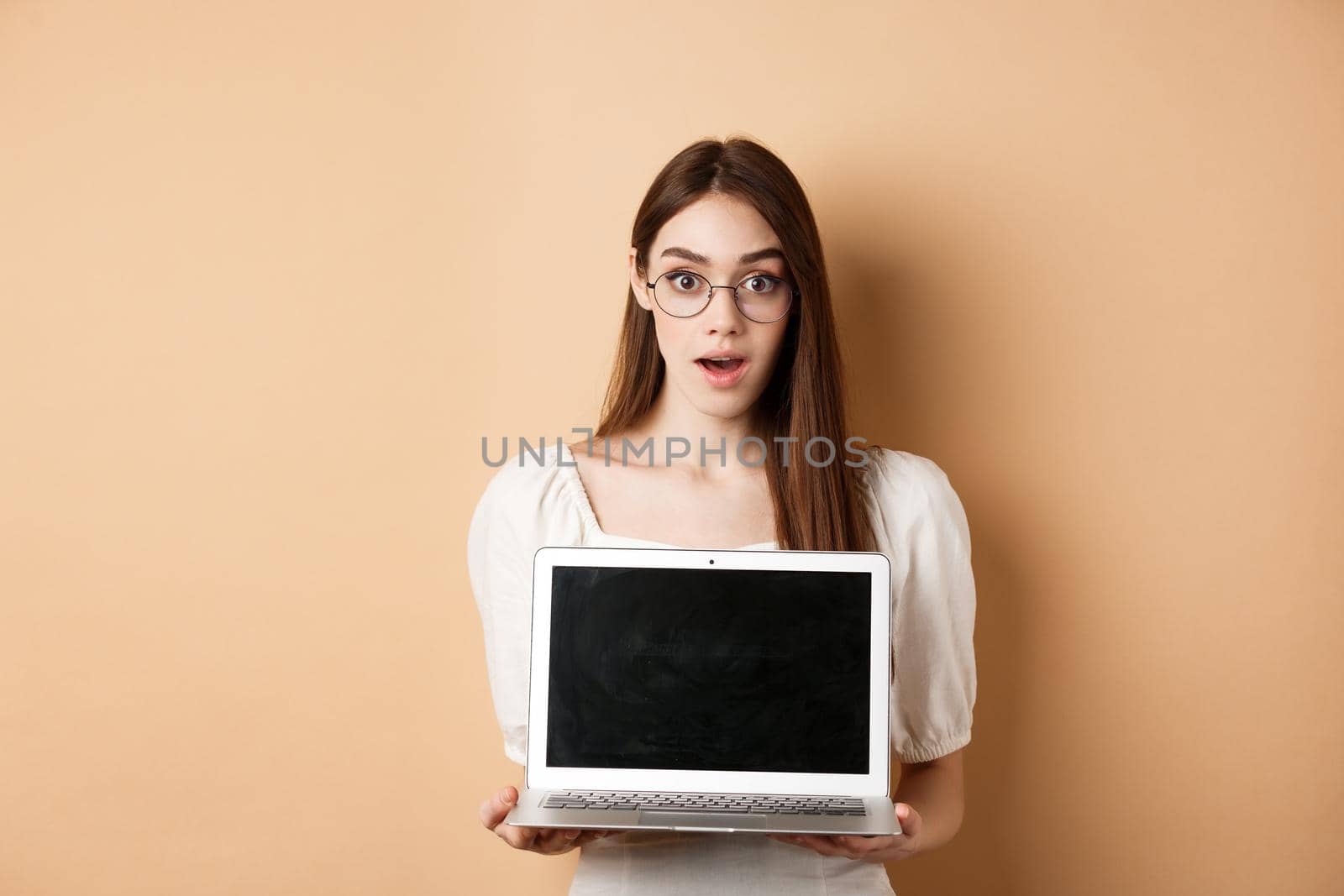 Surprised young woman in glasses showing laptop screen with amazed face, standing on beige background.