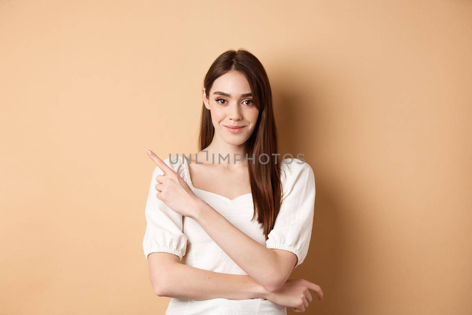 Smiling good-looking girl in white dress pointing aside, showing left logo and staring at camera confident, standing on beige background.