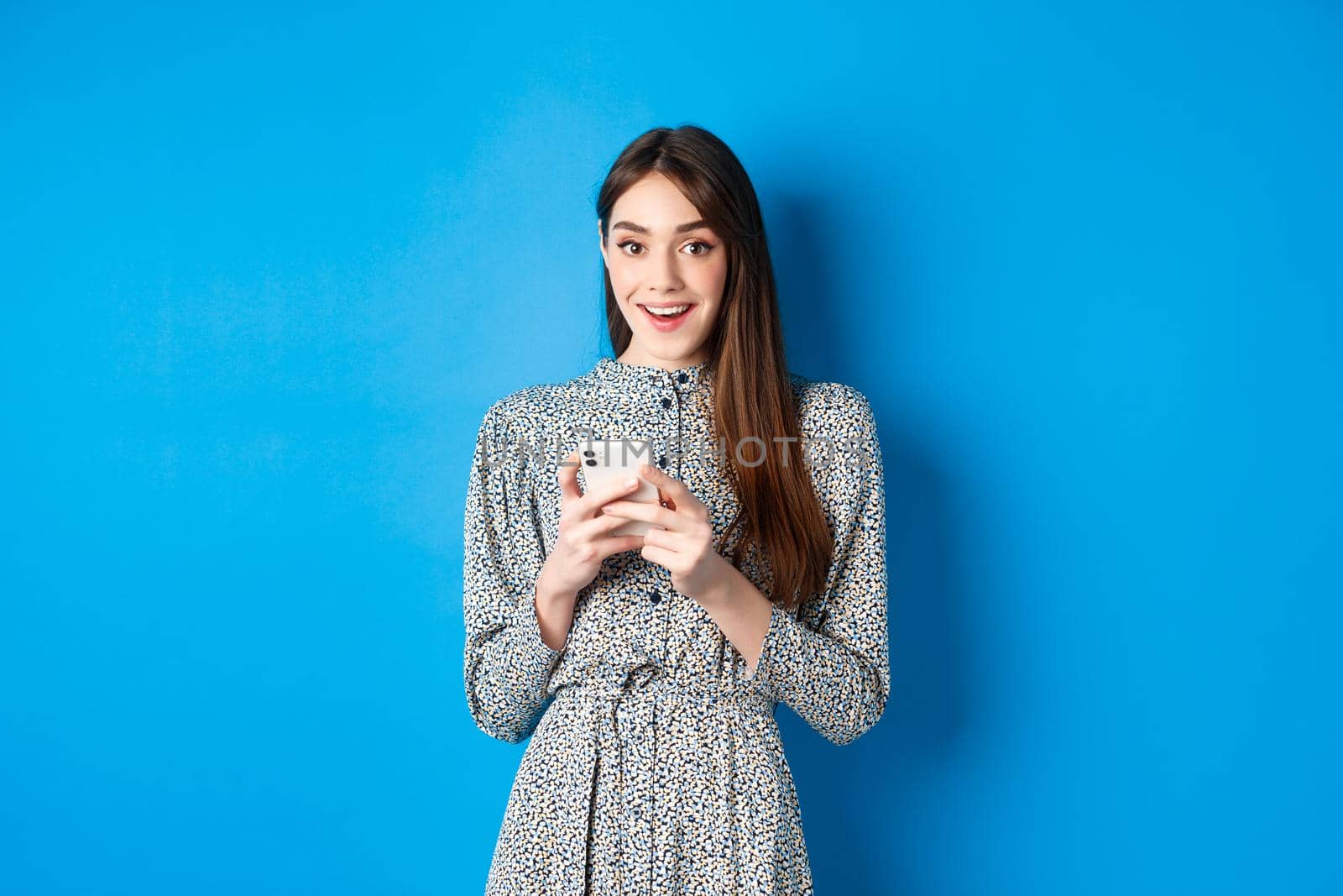 Cute smiling girl with long natural hair, wearing dress, using smartphone and looking happy, standing against blue background.