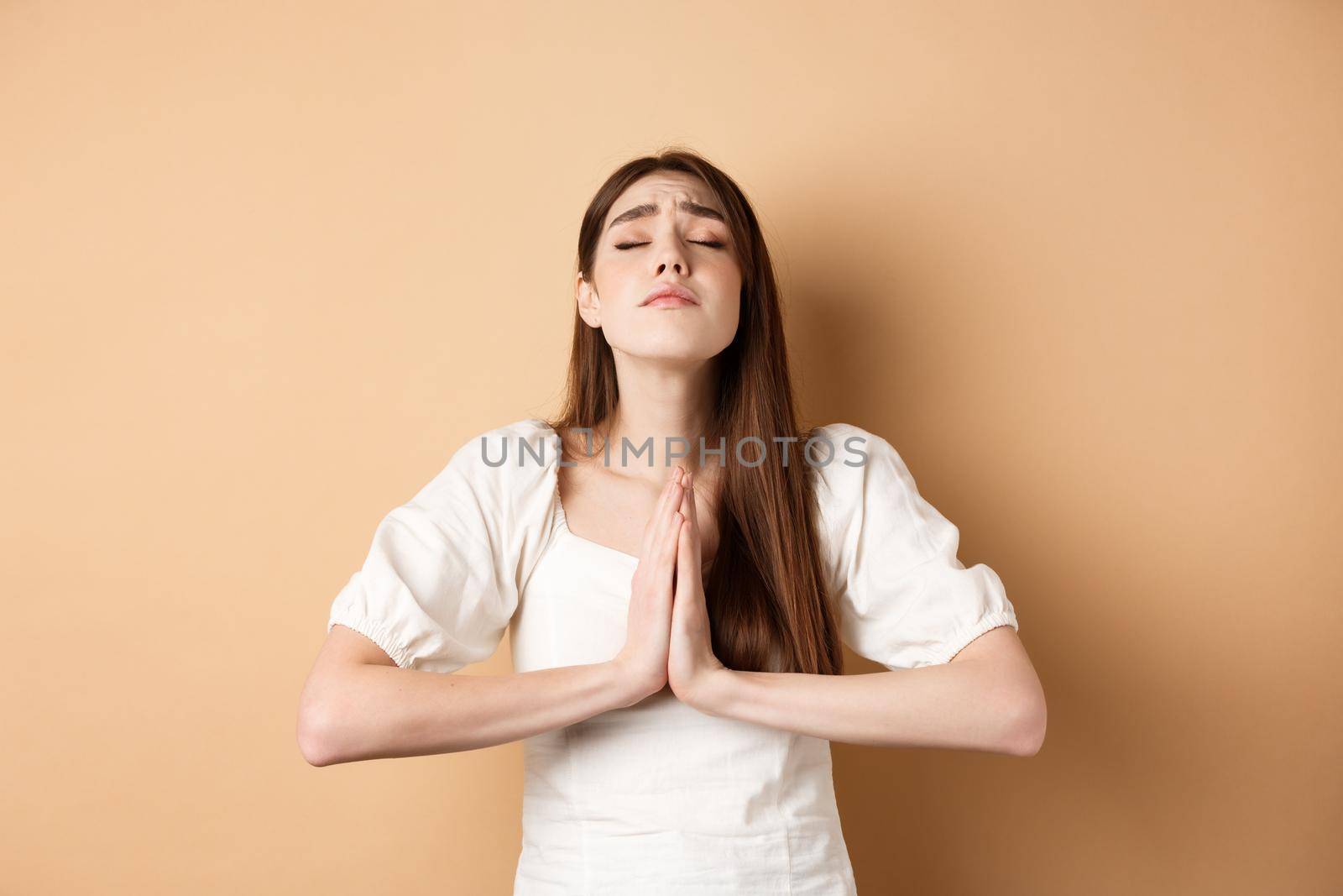 Hopeful woman close eyes and pray god, holding hands in begging gesture and making wish, say please, standing on beige background.