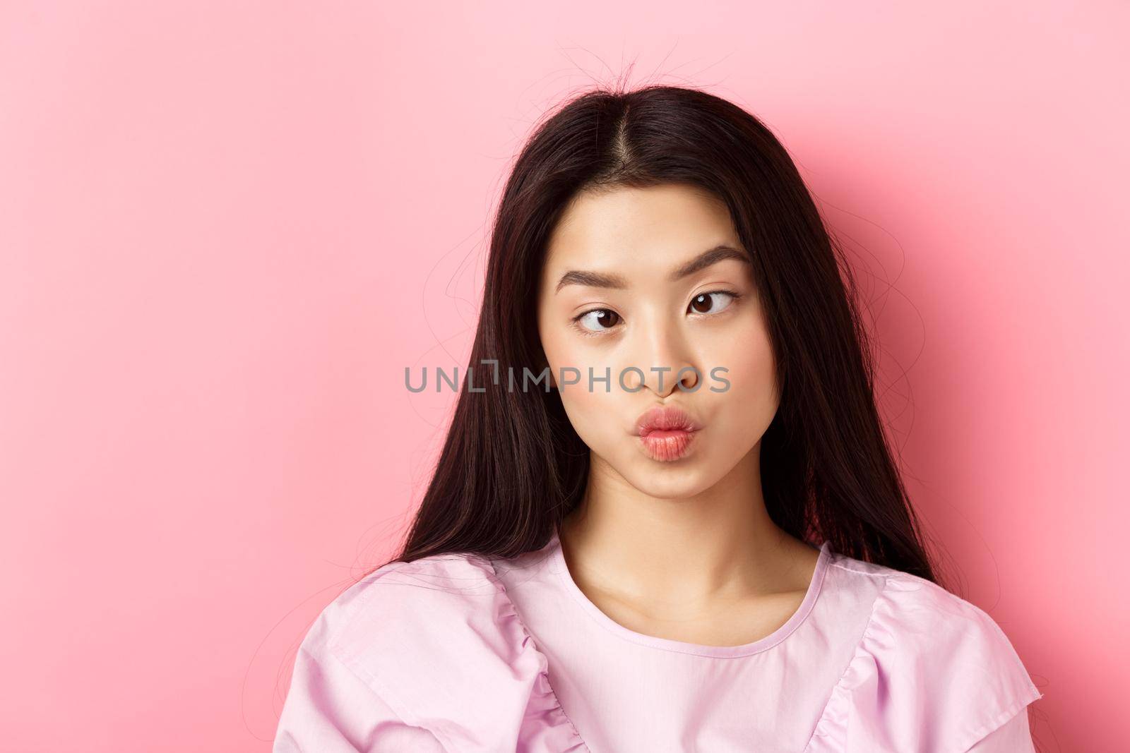 Close-up portrait of funny asian woman squinting eyes and making silly faces, standing against pink background by Benzoix