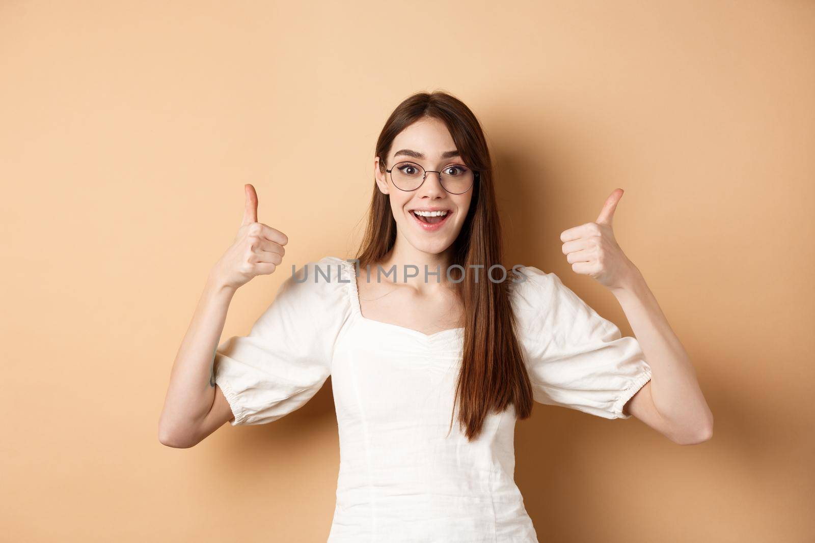 Excited young woman in glasses showing thumbs up and smiling, praising good optic store, approve and like eyewear, standing on beige background by Benzoix