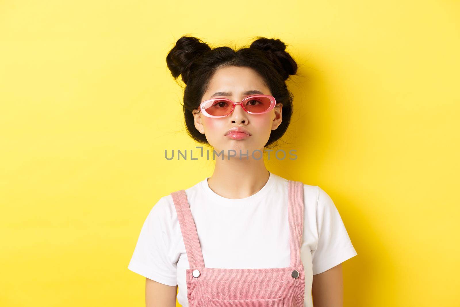 Summer and fashion concept. Bored asian teen girl in sunglasses pouting, standing moody on yellow background.