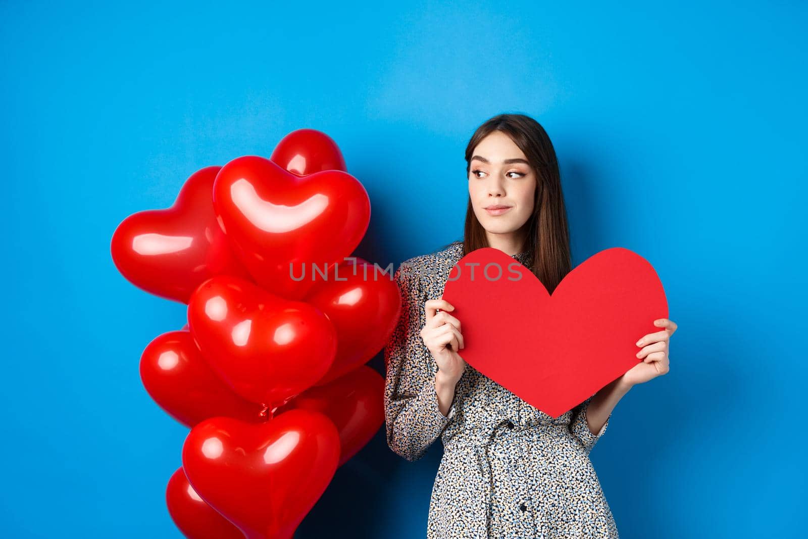 Valentines day. Romantic girl in dress standing near balloons and holding big red heart cutout, dream of something, standing on blue background.