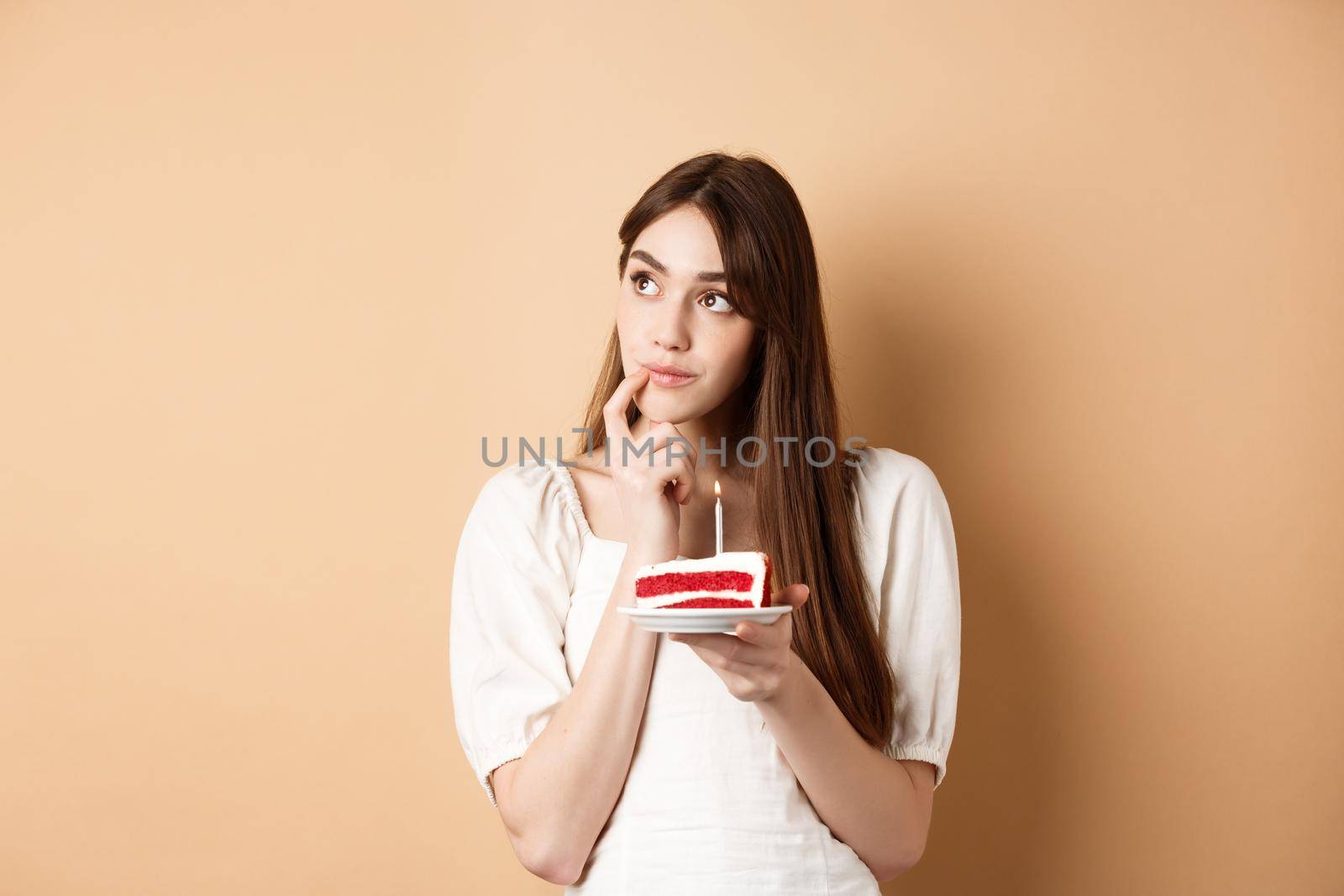 Dreamy birthday girl look aside and thinking of wish, blowing candle on cake, standing on beige background.