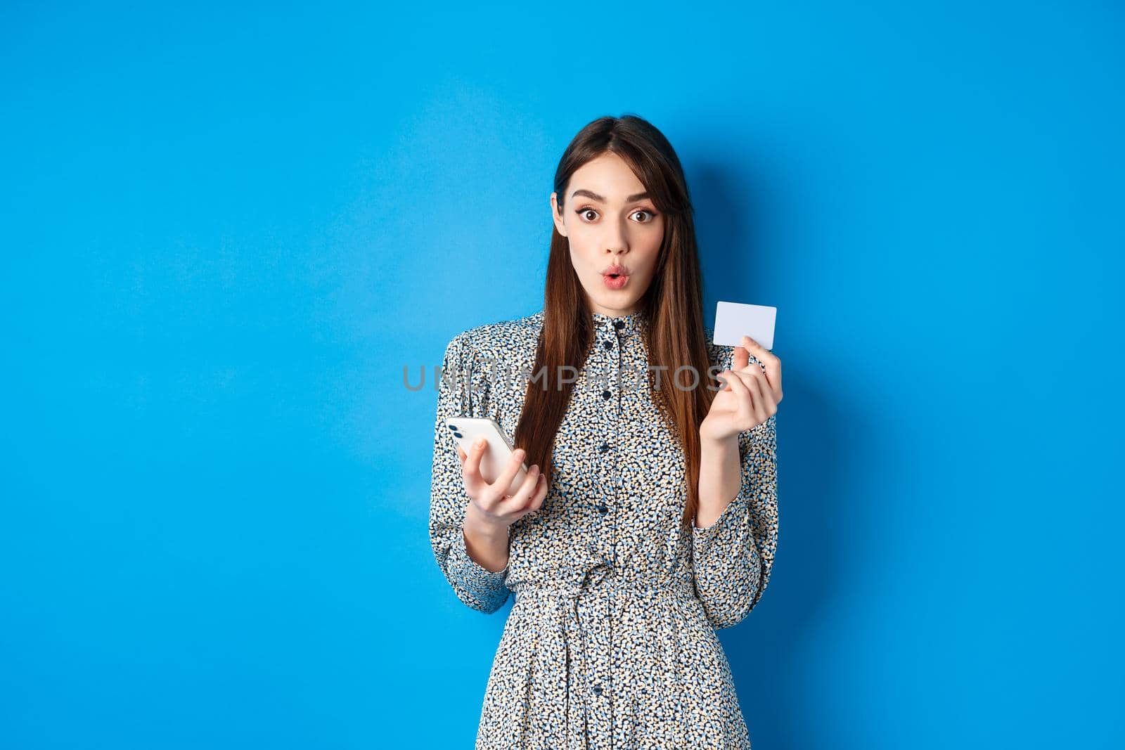 Online shopping. Excited girl paying with credit card on smartphone app, look amused, standing on blue background.