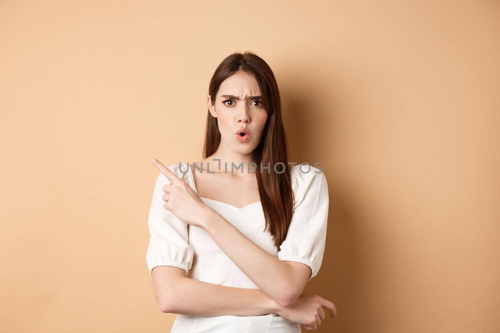 What is that. Shocked and angry woman scolding person, frowning and pointing left with confused face, waiting for explanation, standing on beige background.