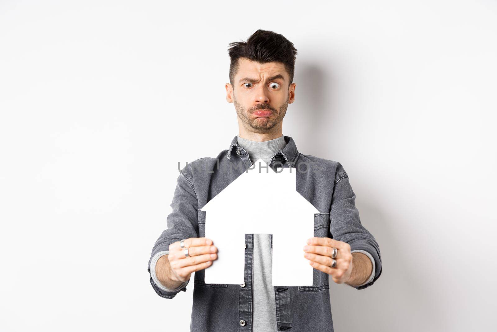 Real estate and insurance concept. Confused guy look at paper house cutout unsure, frowning perplexed, standing on white background.