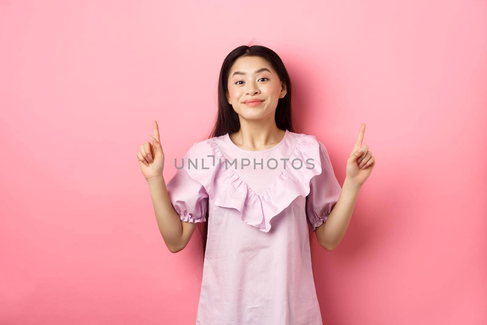Beautiful young asian woman in romantic dress pointing fingers up, smiling and gladly showing advertisement, standing on pink background by Benzoix