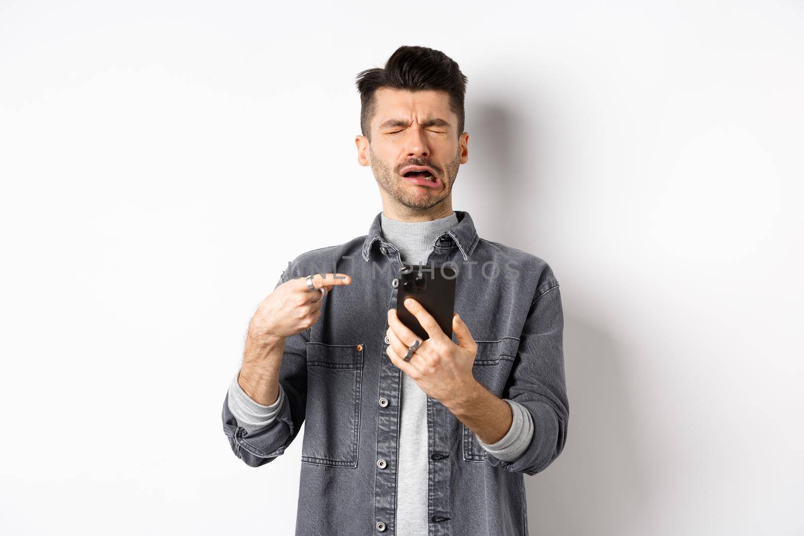 Sad crying guy pointing at smartphone and sobbing, complaining or feeling miserable, standing on white background by Benzoix