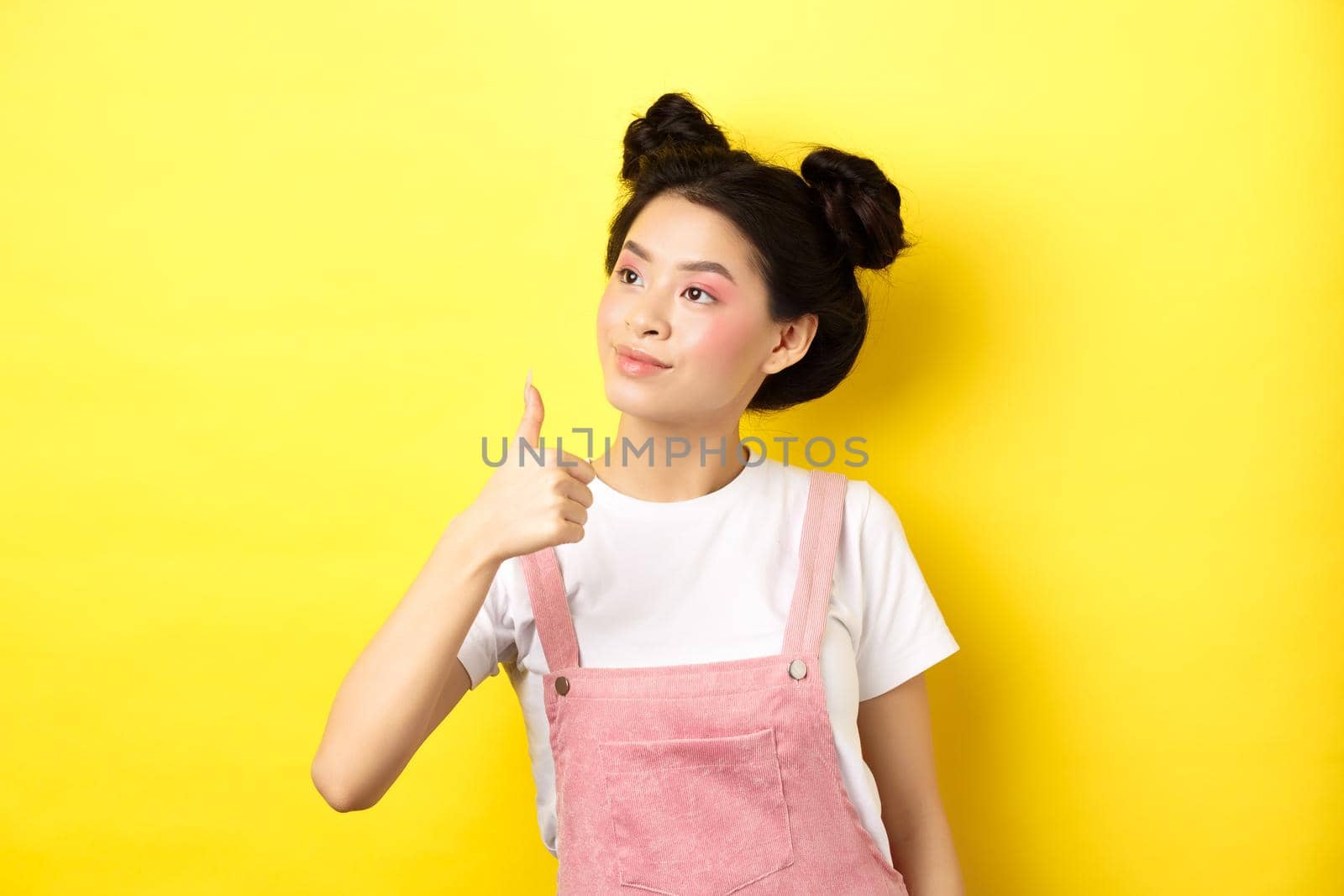Glamour asian girl with pink makeup and summer clothes, looking left at logo with thumb up, approve and praise, standing on yellow background by Benzoix