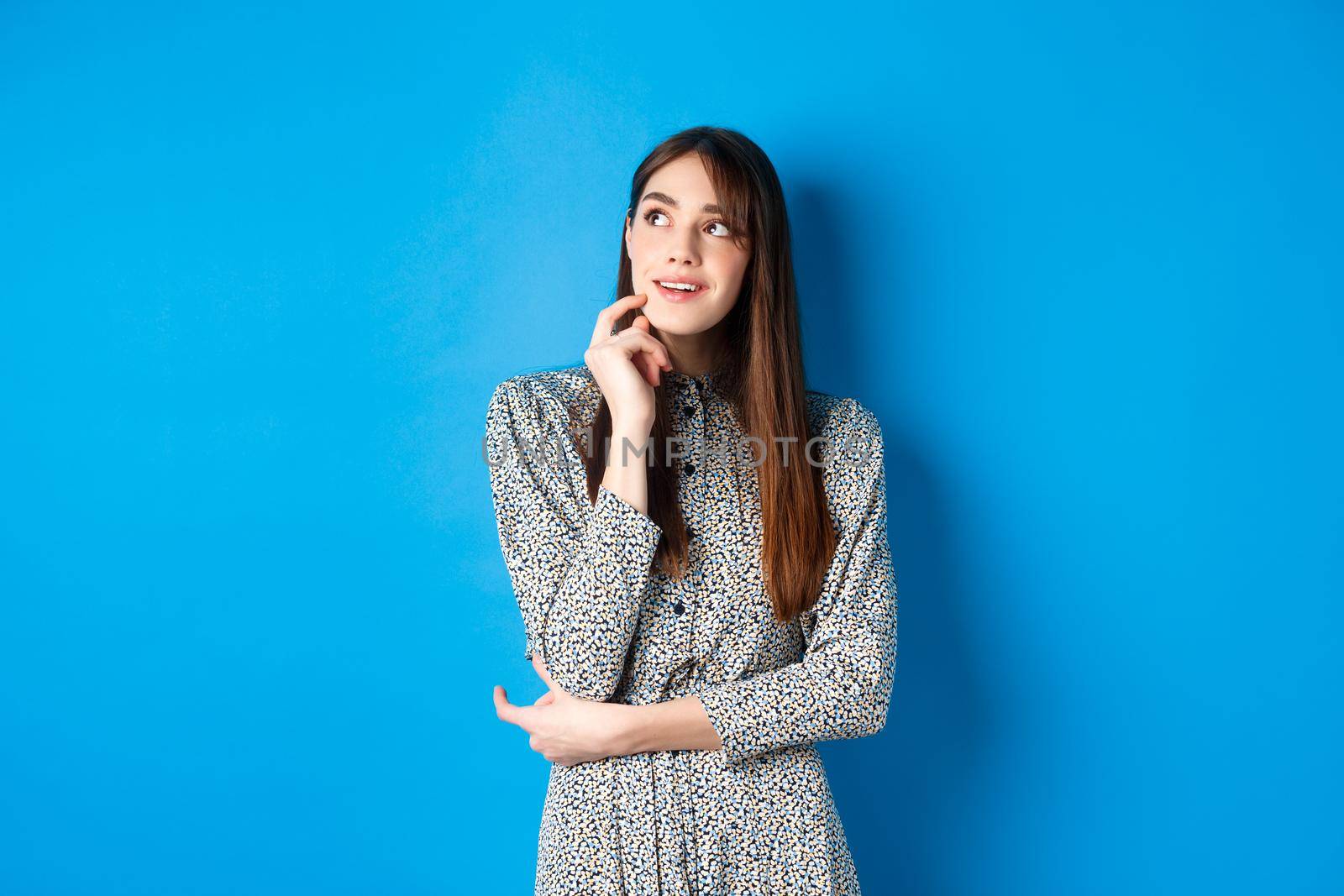 Creative young woman imaging and smiling, looking at upper left logo thoughtful, having an idea, standing on blue background in dress.