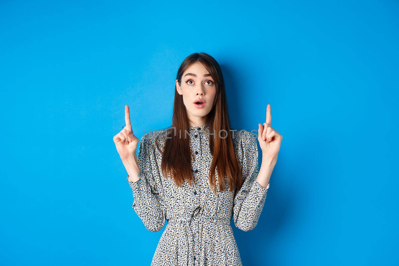 Surprised romantic girl in dress pointing, looking up with opened mouth and fascinated gaze, standing on blue background.