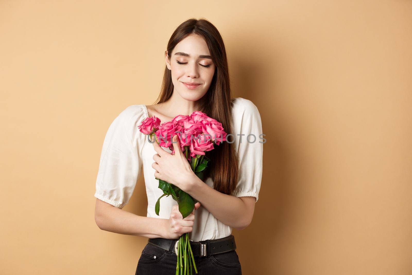 Valentines day. Tender and romantic girl, smell roses and smile with closed eyes. Girlfriend hugging gift flowers from lover, standing on beige background by Benzoix
