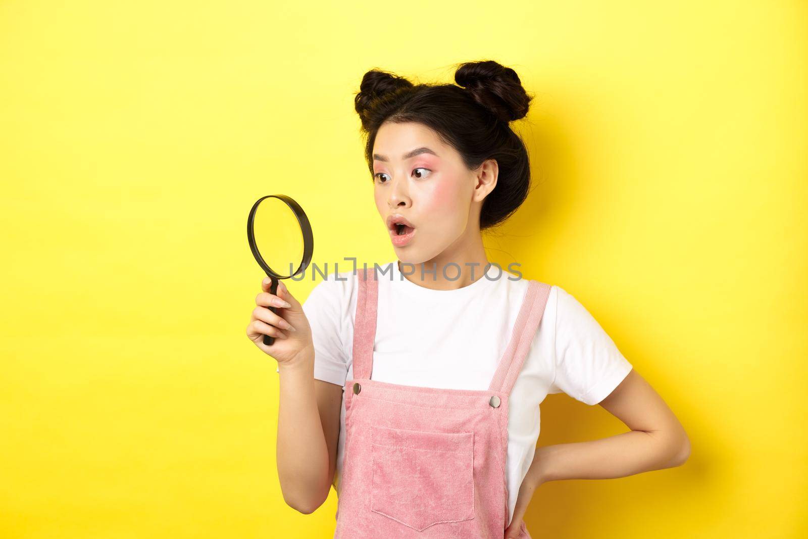 Excited asian woman with bright glamour makeup, look through magnifying glass amazed, staring aside, standing on yellow background.