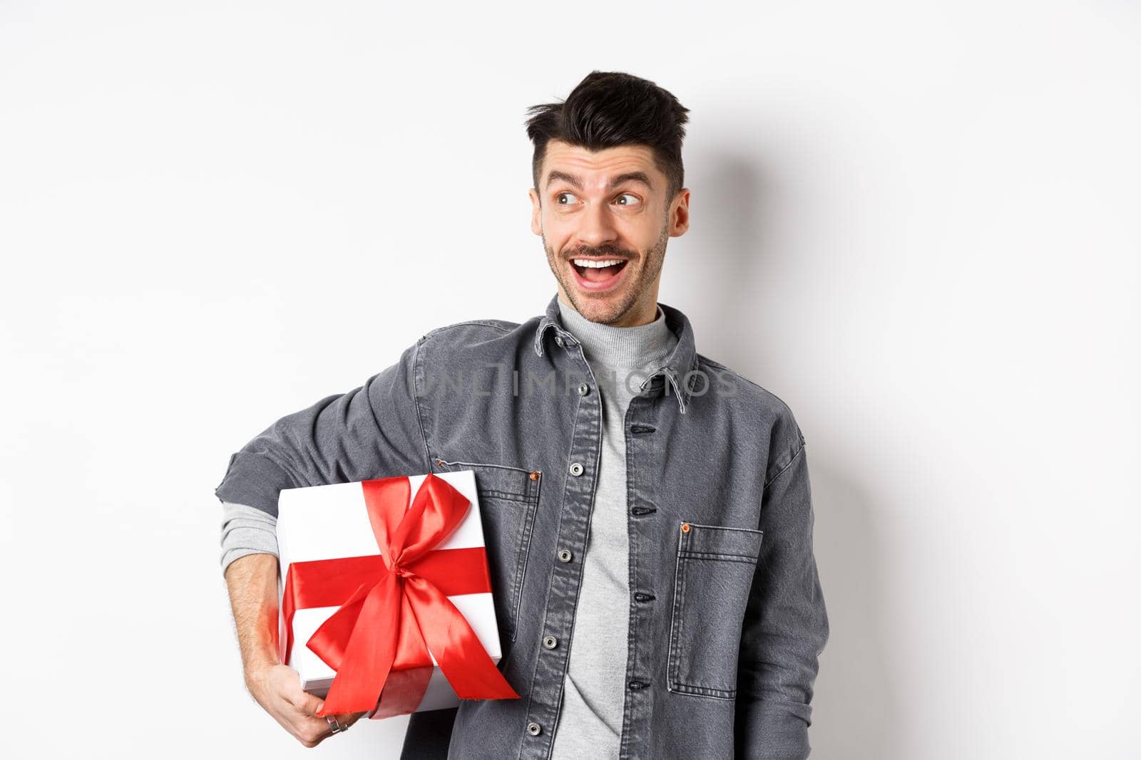 Cheerful handsome guy looking at empty space with amazed smile, checking out special valentines day offer, holding big romantic gift box, standing on white background by Benzoix