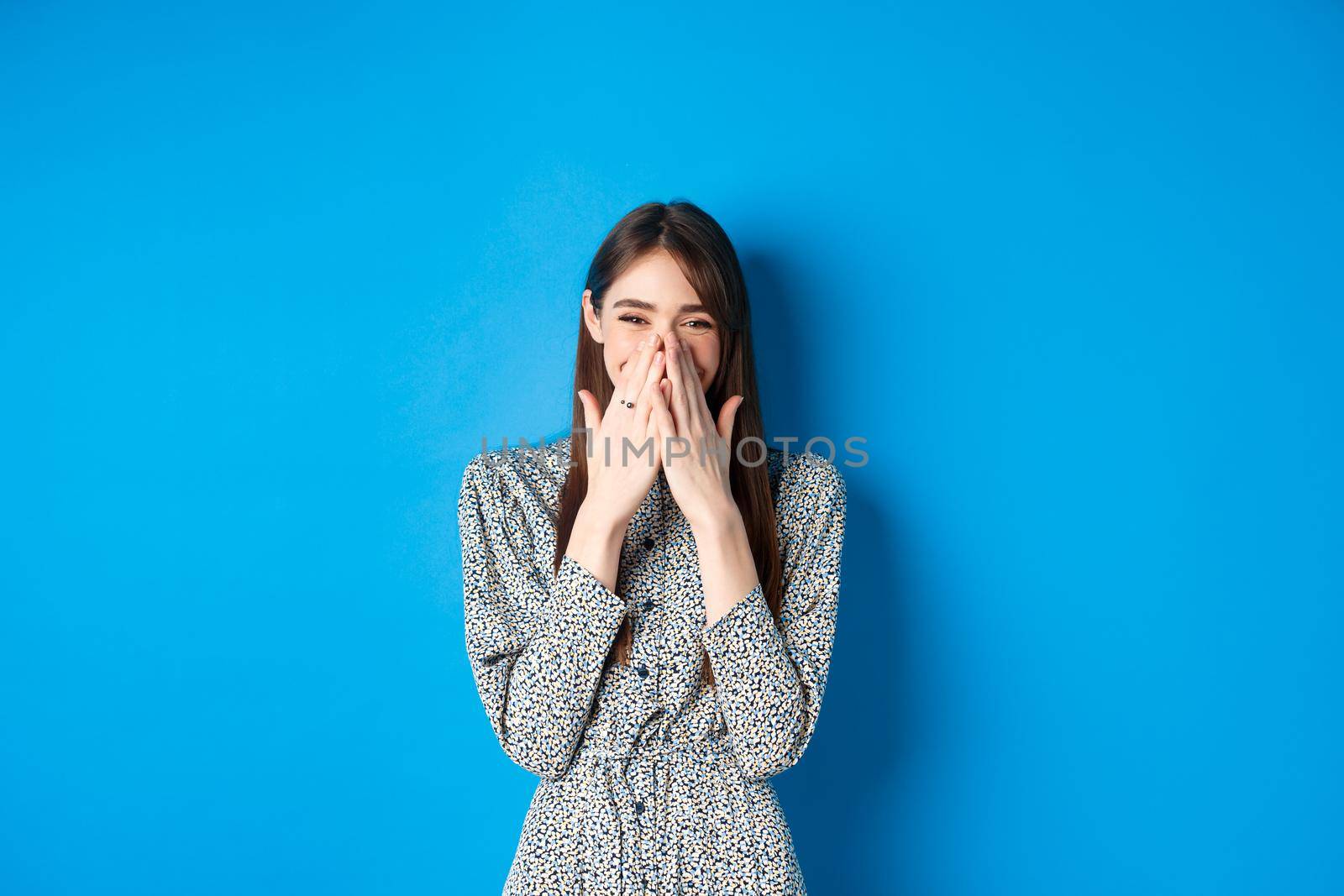 Cheerful caucasina girl in dress laughing and having fun, covering mouth with hands and chuckle over something funny, standing on blue background.