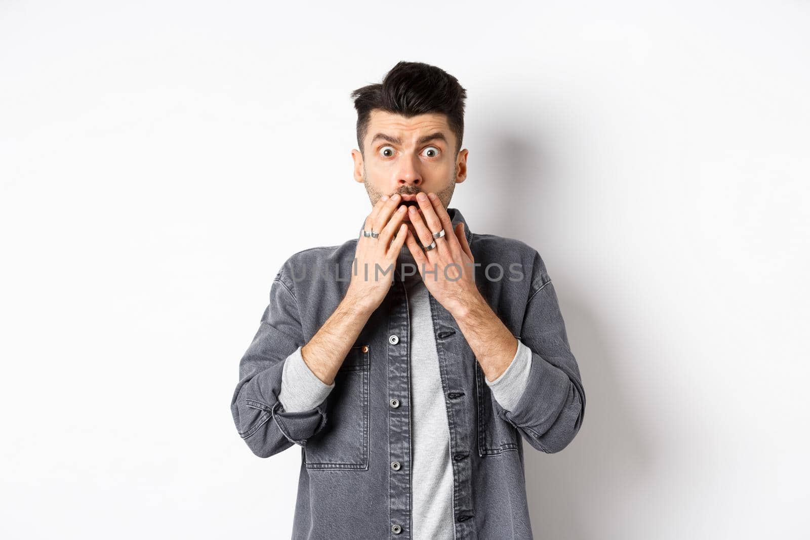 Shocked caucasian guy gasping alarmed, cover mouth with hands and staring at something terrible, witness bad accident, say oh my god, standing on white background.
