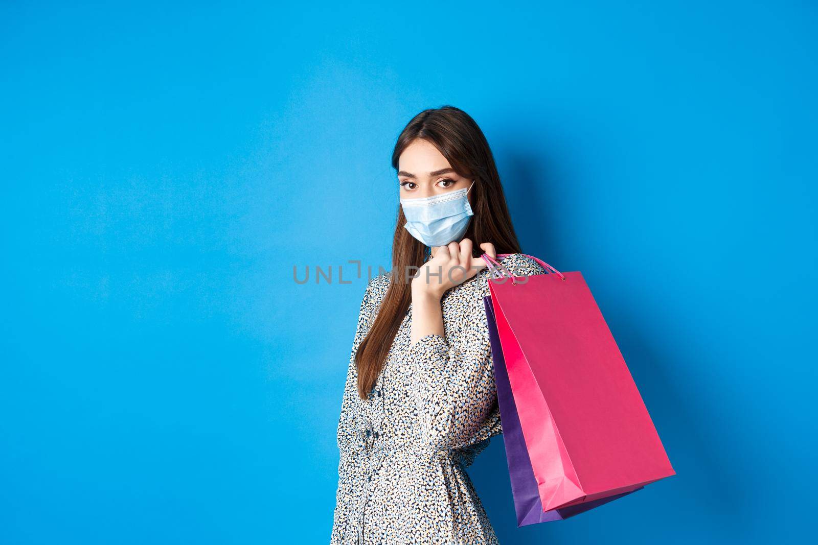 Covid-19, pandemic and lifestyle concept. Elegant woman wear medical mask on shopping, holding bags over shoulder and walking against blue background by Benzoix