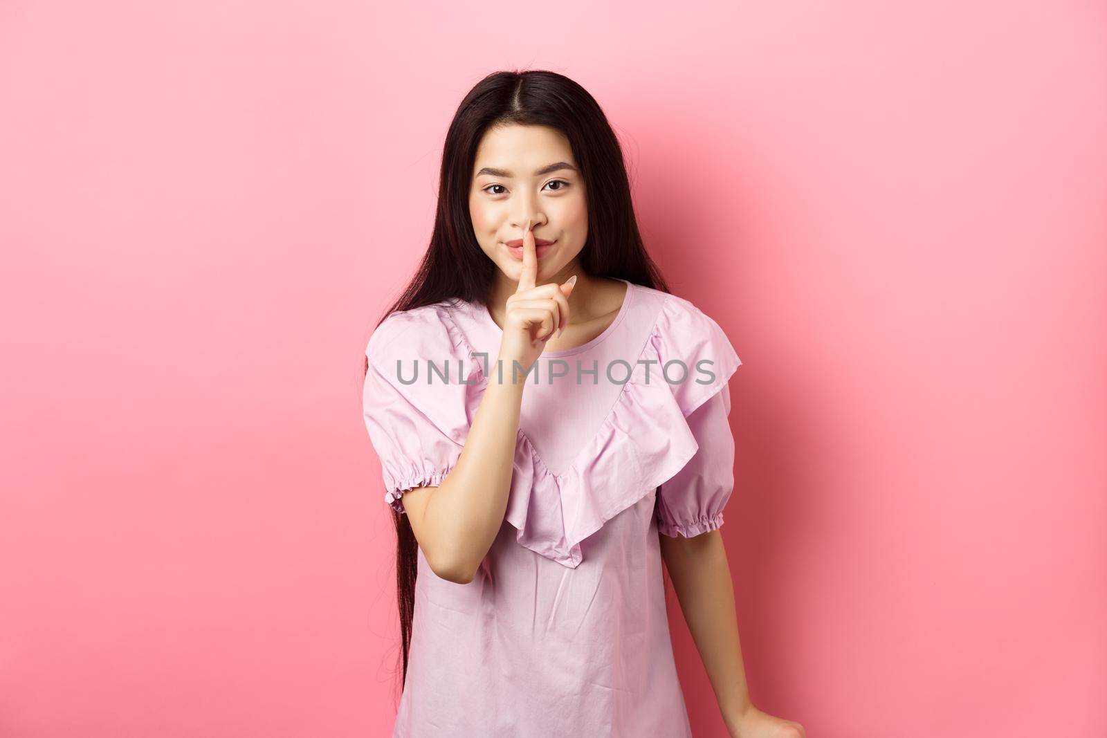 Cute asian girl hiding secret, hushing with finger pressed to lips and smiling, asking to keep quiet, standing in dress on pink background by Benzoix