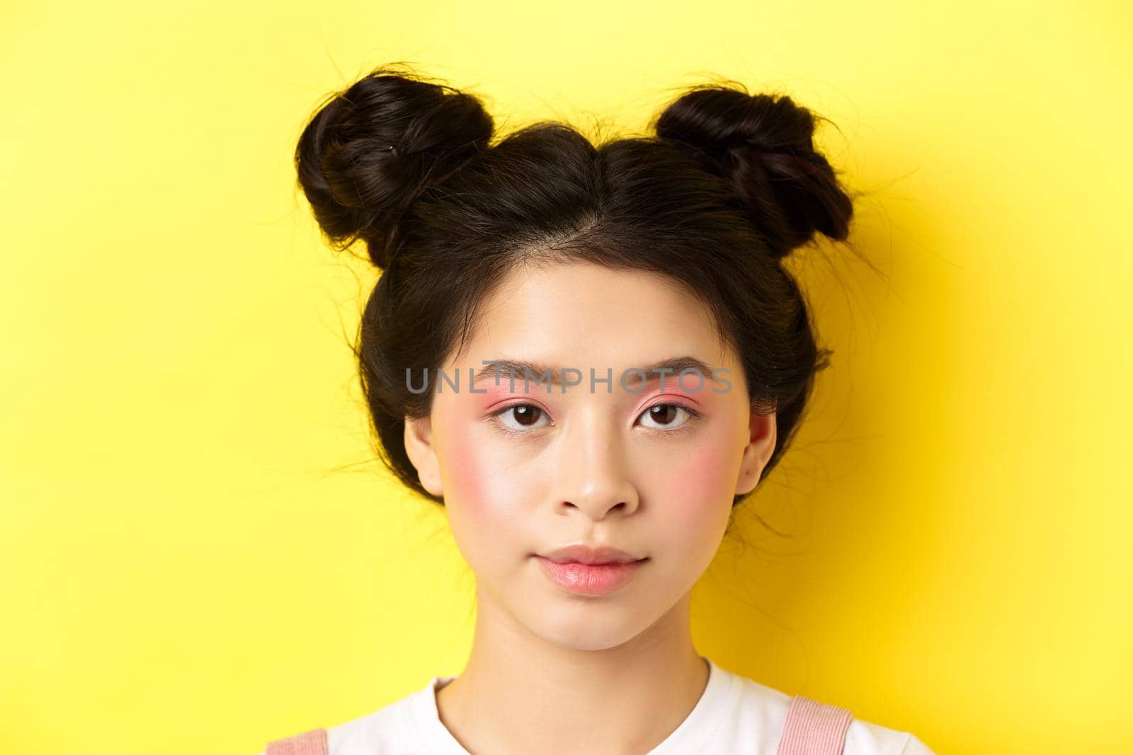 Head portrait of stylish asian girl with bright makeup and hairbuns, looking at camera, standing on yellow background.