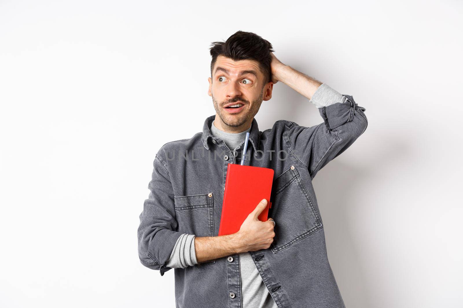 Confused guy scratch head and look aside at logo puzzled, holding red diary or planner, standing against white background.