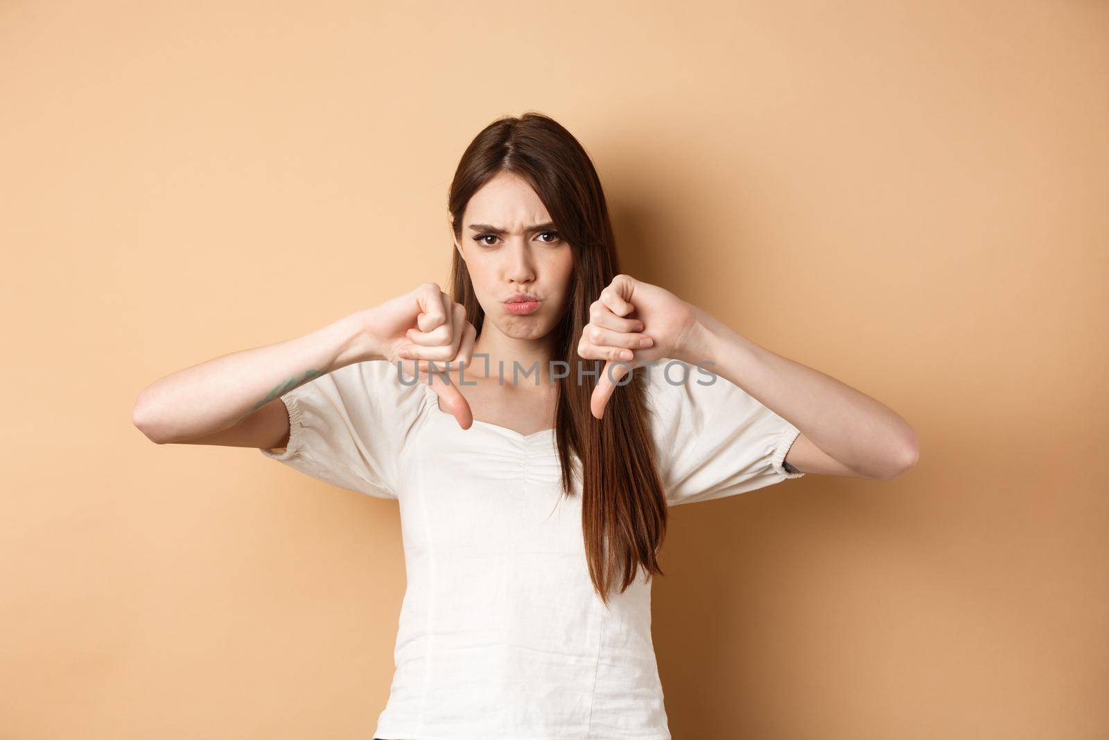 Absolutely no. Disappointed young woman frowning and showing thumbs down, express dislike and negative emotions, bad feedback, standing on beige background.