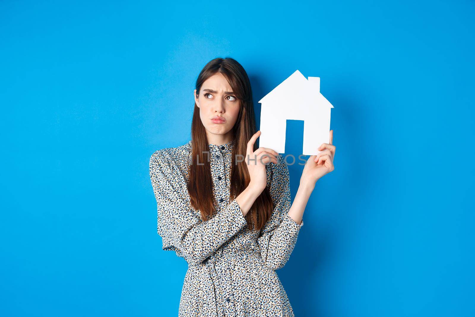 Real estate. Sad frowning girl looking aside with unfair and upset face, showing paper house cutout, standing on blue background.